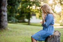 Blonde Woman Sitting On A Tree Stump And Praying In A Garden Under Sunlight