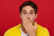 © MarHo - scared handsome man in yellow shirt covering mouth with hand, stares at camera, has bated breath, isolated on red background