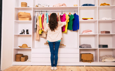 Choosing an outfit. A young girl in a cardigan is standing with her back to the camera, facing a big white compartment wardrobe and trying to choose clothes to wear.