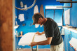 © Natalia Nieves/ADDICTIVE STOCK - Side view of adult man measuring white board while working in small workshop with blue walls and making surf board
