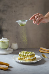 Female hand sprinkling matcha powder over tiramisu cake with green tea or spirulina. Superfoods, healthy nutrition. Confectionery, bakery, cafe menu concept.