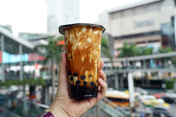 Bubble Milk Tea - Holding a plastic glass of fresh milk with black sugar syrup (Kuromitsu) and hot black pearl (Boba) topped with cream cheese foam on blurred background, Taiwanese style.