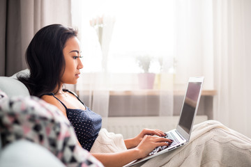 Wall Mural - Relaxed asian girl using laptop sitting on sofa at home