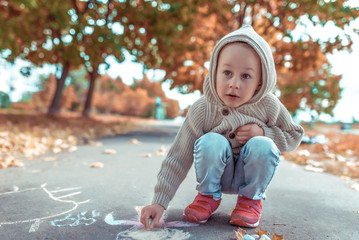 Little boy 3-5 years old, happy draws colorful crayons pavement, creativity park, feel happy. Casual warm clothes with hood. Autumn day. Free space copying text. Background of orange autumn leaves.