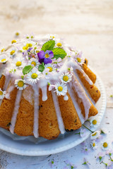 Wall Mural - Easter yeast cake (Babka) covered with icing and decorated with edible flowers on a white plate on a white wooden table, close-up. Traditional Easter cake in Poland