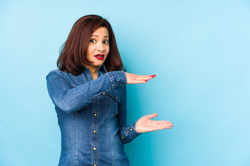 Middle age latin woman isolated on a blue background shocked and amazed holding a copy space between hands.