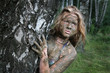 A girl with a mud mask peeps out from behind a birch trunk at an ecological resort. Beautiful blonde