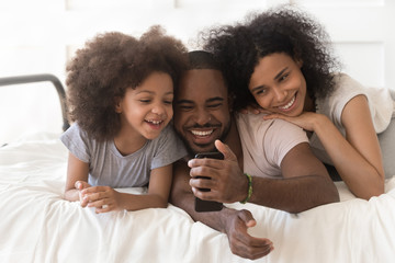 Wall Mural - Happy african parents and daughter lying in bed using smartphone