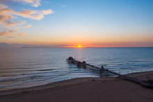Aerial View Of Wooden Pier In Huntington Beach, Orange County In Southern California At Sunset With Waves Crashing Below At Sunset.