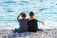 Two Boys Sitting On The Beach And Throwing Rocks Into The Sea