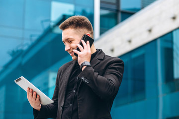 The guy in the black suit on the background of the glass modern business center. confident businessman talking on phone walking in front of modern architecture. Portrait of a smiling businessman