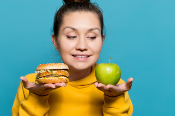 Wall Mural - girl holds an apple and a burger on the palms near her face and smiles, looking at the apple