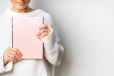 Fototapeta Sypialnia - Close view of woman in white woolen sweater holding a book with empty pink cover in hands. Free space for your mock up of reading book concept background.
