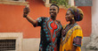 © VAKSMANV - African young stylish and attractive couple in traditional outfits standing together outside and posing to the smartphone camera while taking selfie photo.