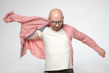 Bald man with glasses putting on a coral-colored shirt, he is preparing for some event and is very serious.