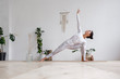 © Rithor - Pretty positive young brunette woman doing Utthita parsvakonasana exercise, Extended Side Angle pose, on mat on floor surrounded by houseplants on white wall. Advertising space. Yoga and pilates