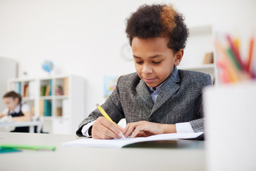 African boy sitting at desk and making notes in his notebook during lesson at school