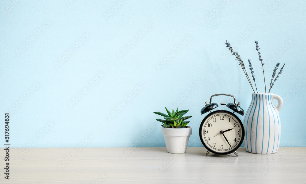 Empty blue wall and desk with alarm clock, home plant and ceramic vase. Copy space - obrazy, fototapety, plakaty 