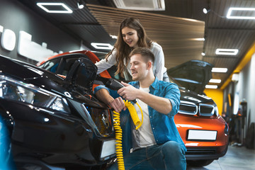 young family couple handsome man shows his beautiful brunette wife how to use charging cable for electric car , looking happy and excited