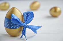 Gold Easter Eggs With Decorative Blue Bow On A Light Background With Space For Text
