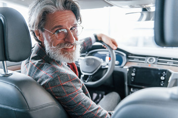 Positive stylish senior man with grey hair and mustache looks back in the modern car