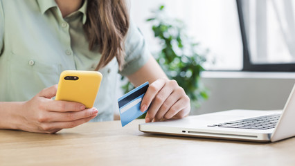 Closeup of young woman hand with credit card and smart phone at home. Businesswoman or entrepreneur working on laptop in office. Online shopping, e-commerce, internet banking, work from home concept