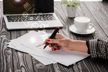 Poster - cropped view of businesswoman holding red marker pen near checklist, illustration, cup and laptop on table, e-commerce concept