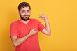 Horizontal shot of male wearing red casual t shirt show his weak biceps muscles, attractive young handsome unshaven man posing isolated over yellow wall background, needs some sport training.