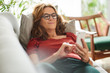 © marvent - Smiling mature woman lying on her couch using a cellphone