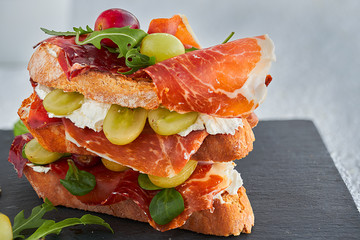 Wall Mural - Front and close-up view of an Iberian ham sandwich with cream cheese, white and purple grapes, canons, arugula, capers and honey on a black background.