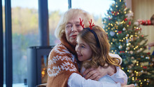 Happy Grandma Holding Her Little Granddaughter On Lap Embracing Together Near Christmas Tree. Cute Adorable Small Girl Wearing Xmas Reindeer Horns. Family Portrait.