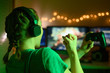 © Anton - The girl emotionally plays video games with a headset, and a joystick, controller, gamepad. On the computer online with her friends. Green creative light