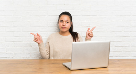 Young teenager Asian girl with a laptop in a table pointing to the laterals having doubts