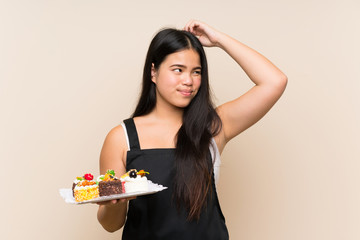 Wall Mural - Young teenager Asian girl holding lots of different mini cakes over isolated background having doubts and with confuse face expression