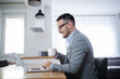© dusanpetkovic1 - Side view of handsome unshaven caucasian businessman in suit and with eyeglasses sitting at dining table, using laptop and drinking morning coffee. Home interior.