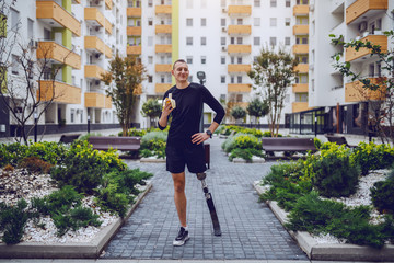 Wall Mural - Full length of attractive caucasian sportsman with artificial leg standing in park surrounded by buildings and eating banana.
