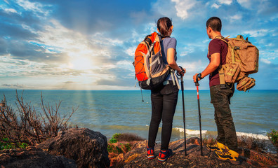 young asian couple hikers climbing up on the peak of mountain near ocean. two tourists with backpack