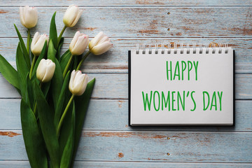 Wall Mural - Bouquet of white flowers of tulips on the background of blue boards with a notebook with lettering Happy Women's Day