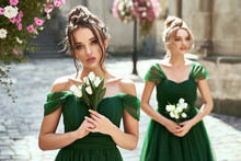 Two beautiful bridesmaids girls blonde and brunette ladies wearing elegant full length off-the-shoulder green chiffon bridesmaid dress and holding bouquets. European old town location for wedding day