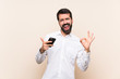 © luismolinero - Young man with beard holding a mobile showing ok sign and thumb up gesture