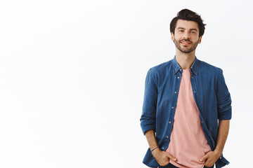 Wall Mural - Cheerful, bearded handsome young man with messy hairstyle, standing relaxed in casual pose with hands in pockets, smiling, ordinary guy shopping in mall, buy gifts. Male look camera enthusiastic