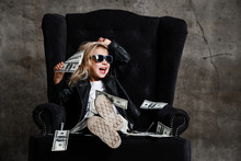 Happy Screaming Loud Laughing Kid Girl Is Sitting In Big Plush Armchair Holding A Wad Of Cash Dollars, Throwing Money