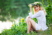 Young Beautiful Blond Woman In White Mini Dress And Floral Wreath Sitting Near Lake And Smiling On Summer Day