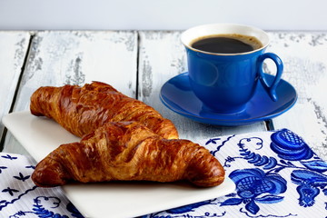 Cup and two croissants on a white plate is on the table of gray boards.