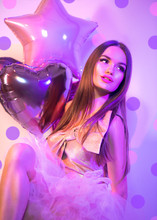 Beauty Fashion Model Party Valentine's Day Girl With Heart Shaped Air Balloons, Sitting On Chair, Laughing In Neon Light Holiday Celebration. Beautiful Young Pin Up Brunette Woman Full Length Portrait