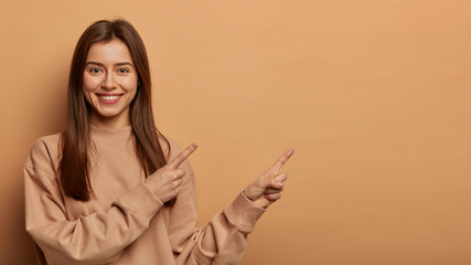 Wall Mural - Look, advertise here! Dark haired pretty woman with appealing smile gives positive attitude towards good deal, expresses her recommendation while pointing on right side, wears brown sweater.