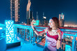 A girl blogger in a rooftop bar takes a selfie photo against the background of a night city with skyscrapers