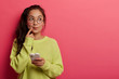 © WHstudio Leushin N - Photo of pensive surprised woman keeps index finger near lips, looks aside, thinks about message content, holds mobile phone, wears transparent glasses and green sweater, isolated on pink wall