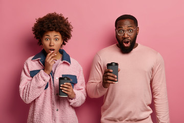 Wall Mural - Isolated shot of stupefied dark skinned young female and male drink takeaway coffee, have shocked expressions, hear unbelievable news, stand next to each other, isolated over pink pastel wall