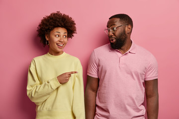 Wall Mural - Delighted curly haired young woman indicates index finger on boyfriend, have cheerful conversation, dressed casually, stand closely, isolated on pink background. Look at my handsome husband.